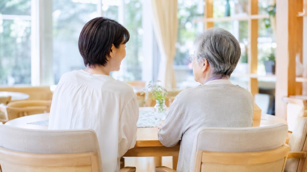 明るい介護施設のラウンジで、入居者とご家族が向き合って談笑する様子(イメージ)